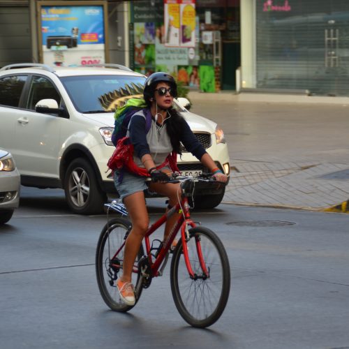 mujer en bicicleta roja en la calle, de fondo dos automóviles